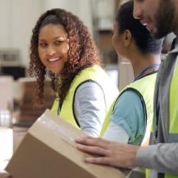 one young black man and two young black women working at a packaging facility. each is wearing a yellow safety vest.